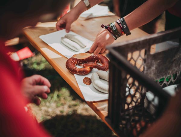 Eine Bretzel wird beim Frühschoppen über den Tresen gegeben. Der Verkäufer trägt viele Festivalbändchen